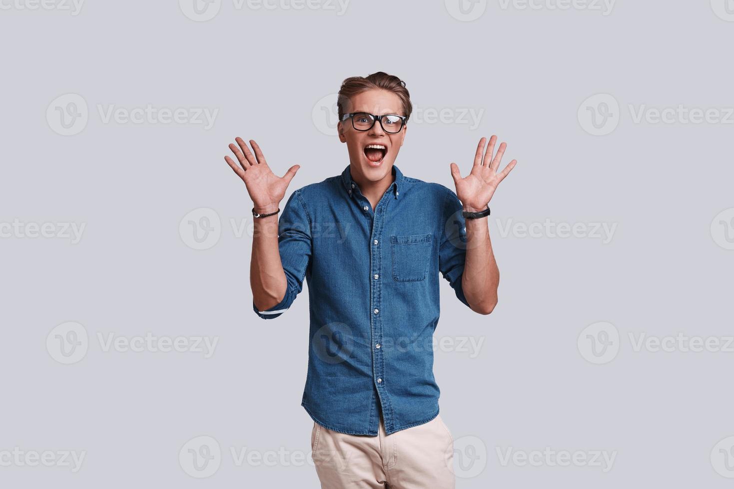 Feeling surprised.  Handsome young man gesturing and keeping mouth open while standing against grey background photo