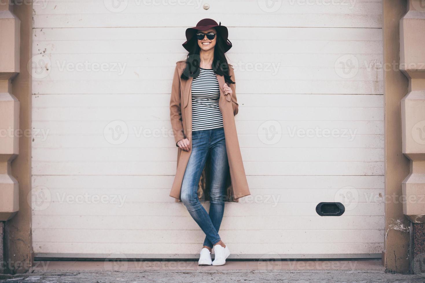 elegante y alegre. toda la longitud de una hermosa joven con gafas de sol mirando a la cámara con una sonrisa mientras está de pie al aire libre foto