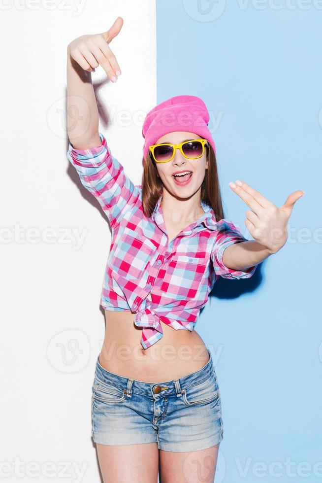 Her energy charges. Playful young woman in headwear and glasses gesturing and looking at camera while standing against colorful background photo