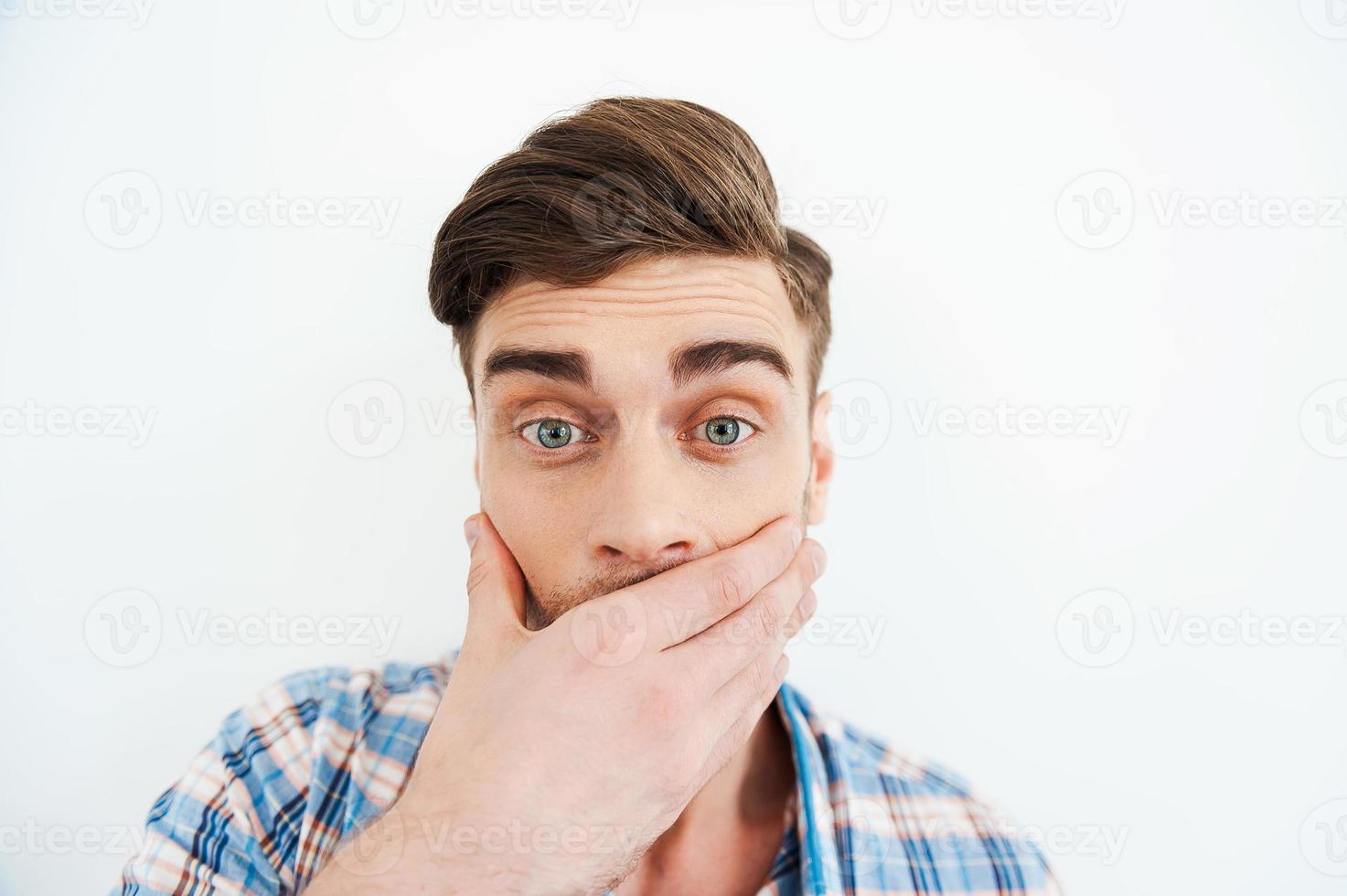 No way Shocked young man covering mouth with hand and looking at camera while standing against white background photo