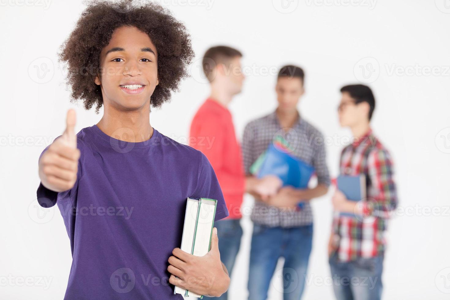 I love studying Cheerful African teenage boy holding books and gesturing while his friends standing on background photo