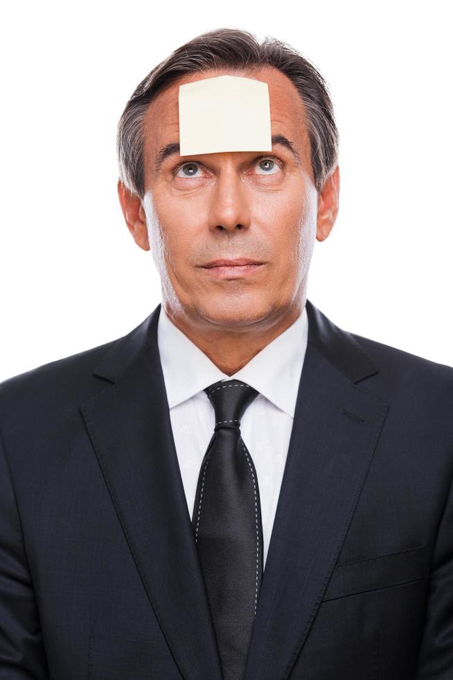 Reminder. Portrait of frustrated mature man in formalwear and adhesive note on his forehead standing against white background photo