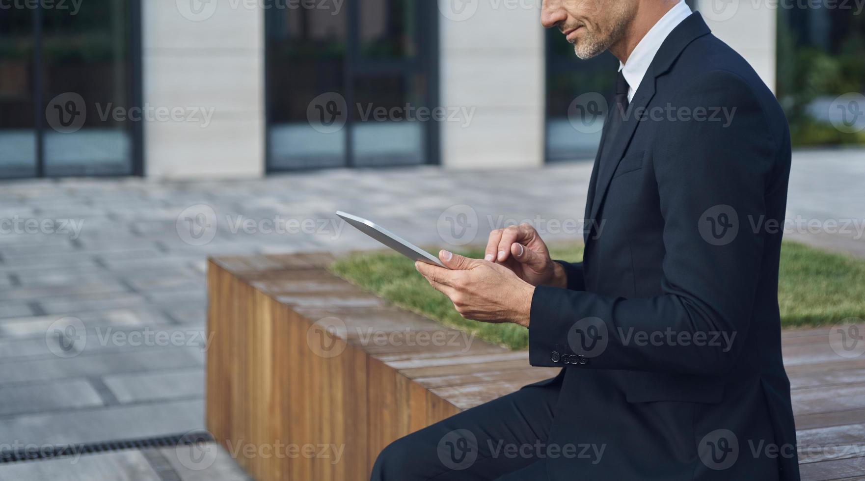 Confident mature businessman using digital tablet while sitting outdoors near office building photo
