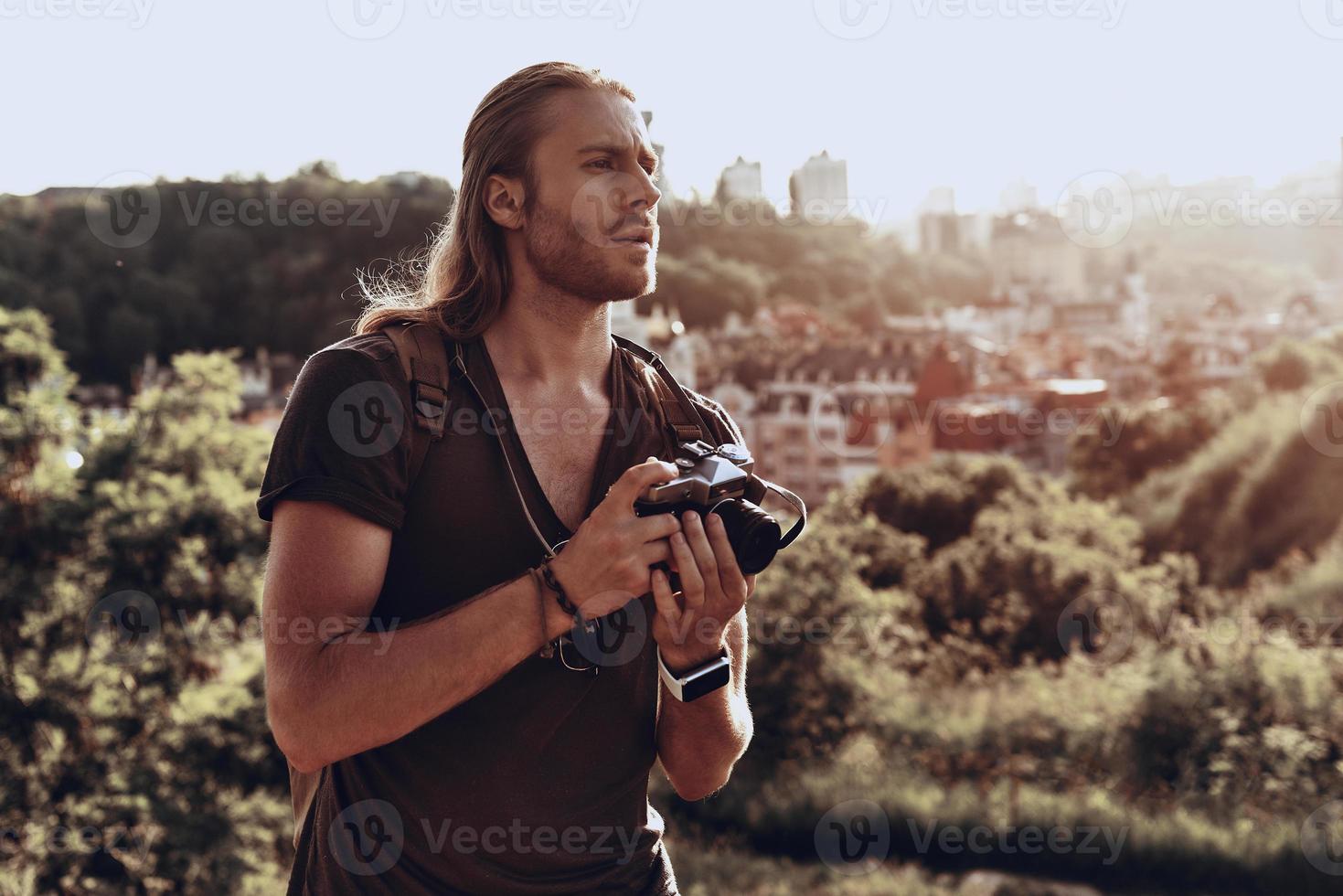 Looking for perfect composition. Young man in casual clothing photographing the view while standing on the hill outdoors photo