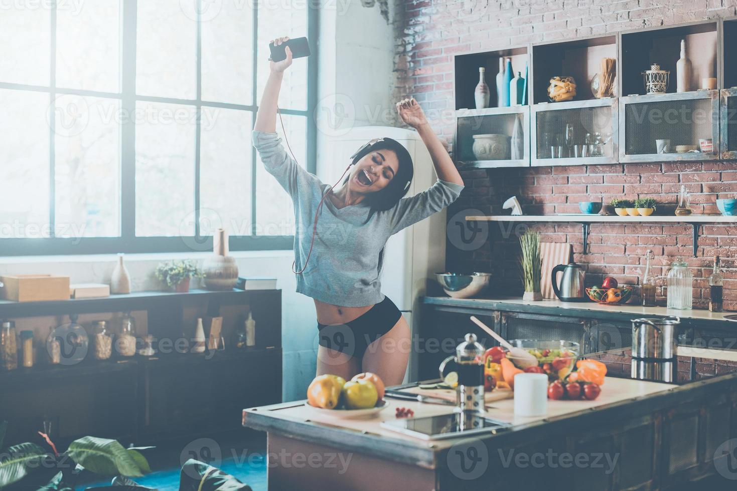 Enjoying new great day. Beautiful young mixed race woman in headphones dancing while standing in kitchen at home photo