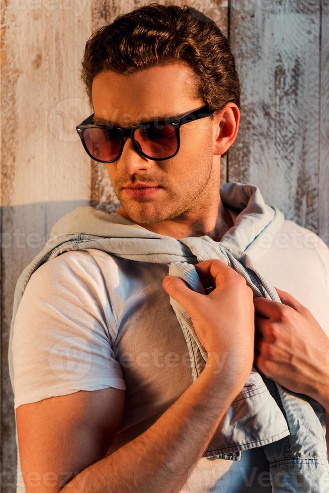 haciendo su estilo. apuesto joven con gafas de sol ajustando su camisa mientras está de pie contra la pared de madera foto