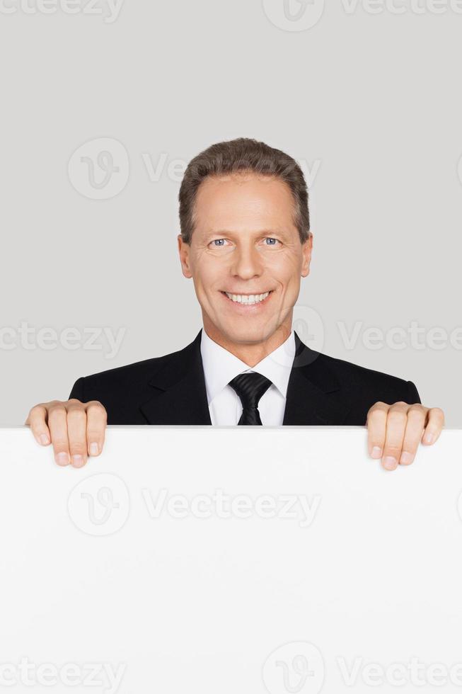 sosteniendo espacio para su texto. hombre maduro alegre en ropa formal mirando fuera del espacio de copia y sonriendo mientras está aislado en fondo gris foto