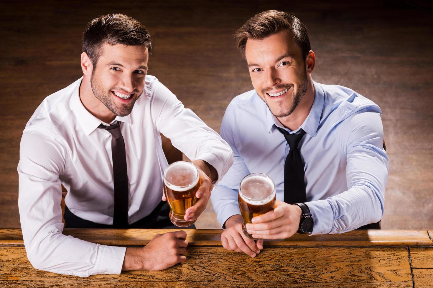 relajante en la noche del viernes. vista superior de dos jóvenes alegres con camisa y corbata sosteniendo vasos con cerveza y sonriendo mientras se sientan en el mostrador del bar foto