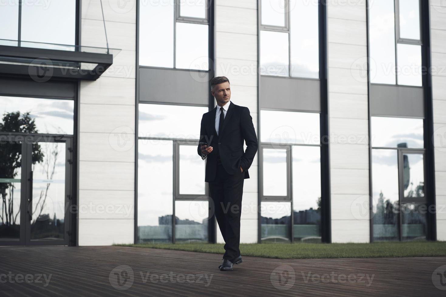 Confident mature businessman holding mobile phone while walking in front of office building photo