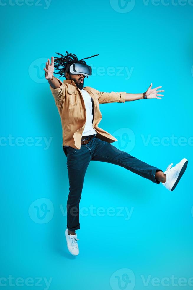 Full length of playful young African man wearing virtual reality glasses while hovering against blue background photo