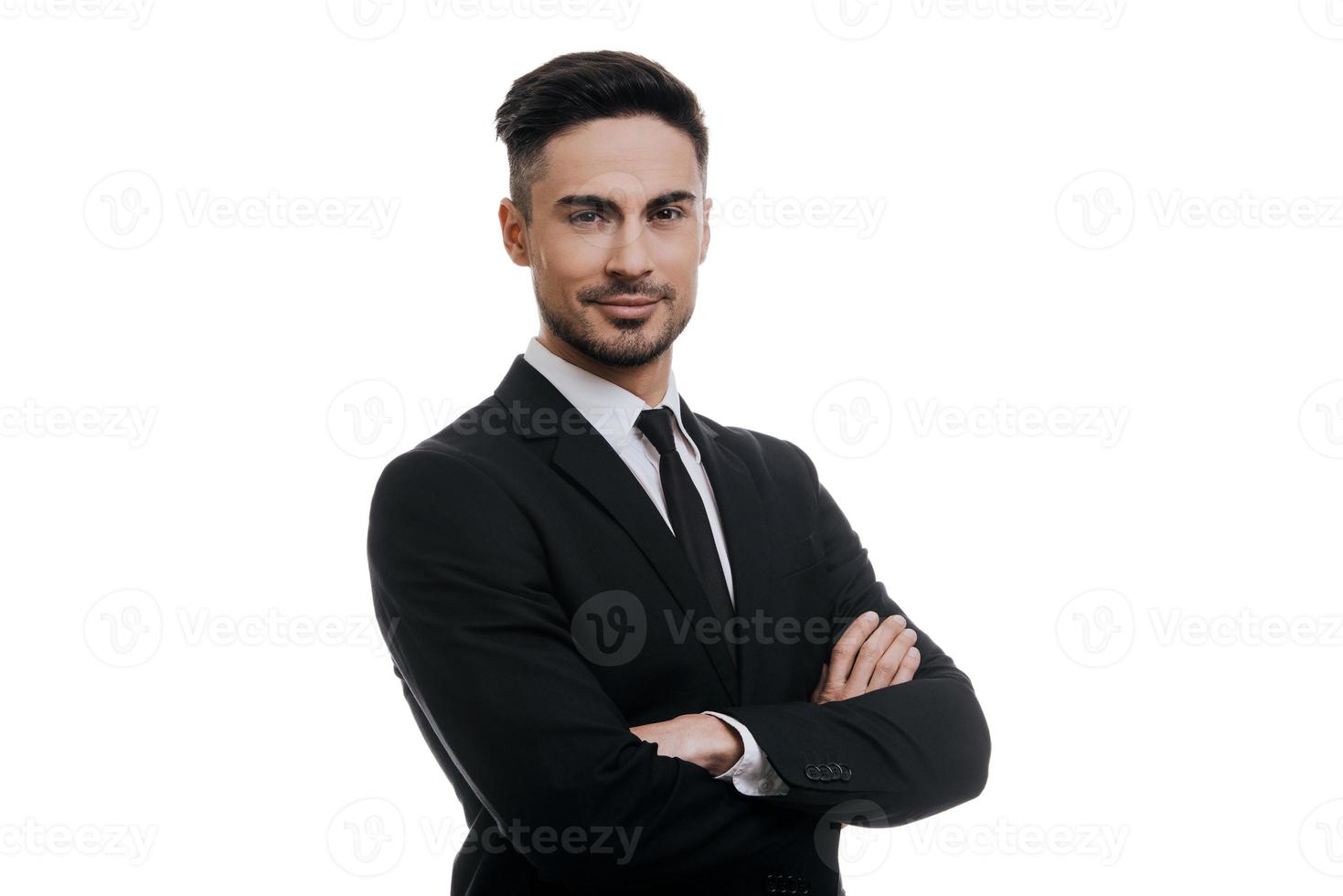 Portrait of elegance.  Handsome young smiling man in full suit looking at camera and keeping arms crossed while standing against white background photo