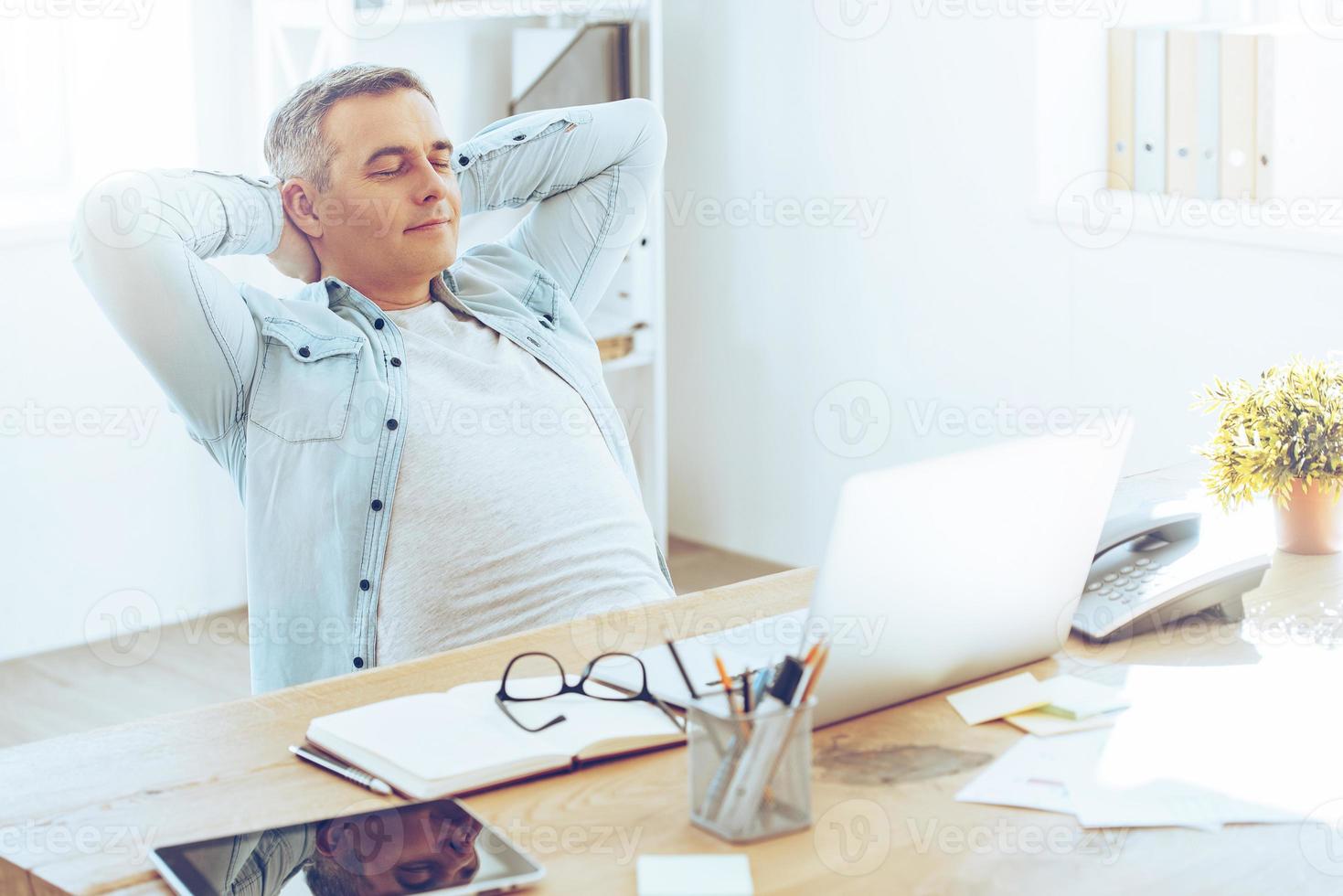 Taking a minute break. Happy mature man looking relaxed and holding hands behind head while sitting at his working place photo