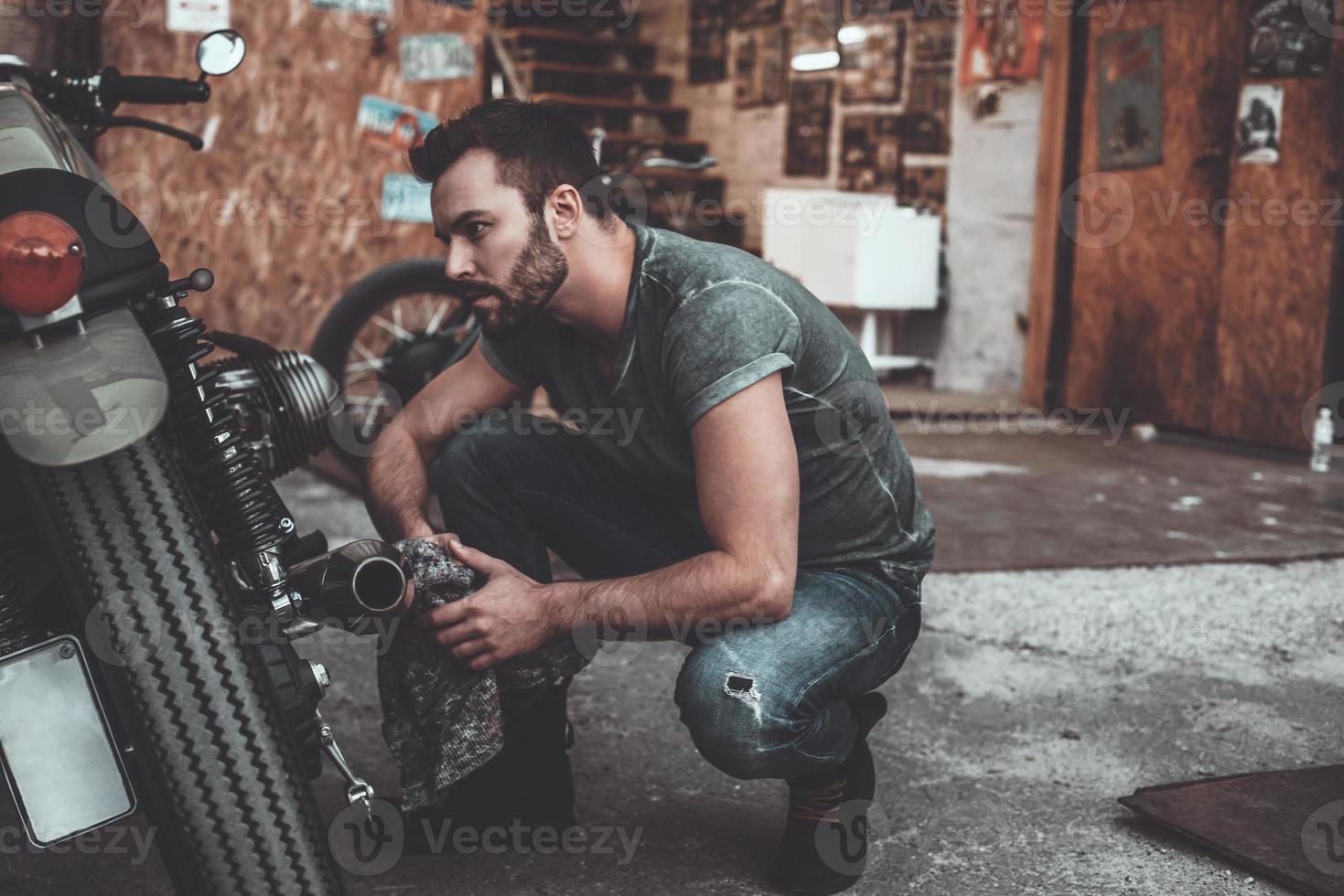 What seems to be a problem Confident young man holding rag and looking at motorcycle while crouching near it with garage in the background photo