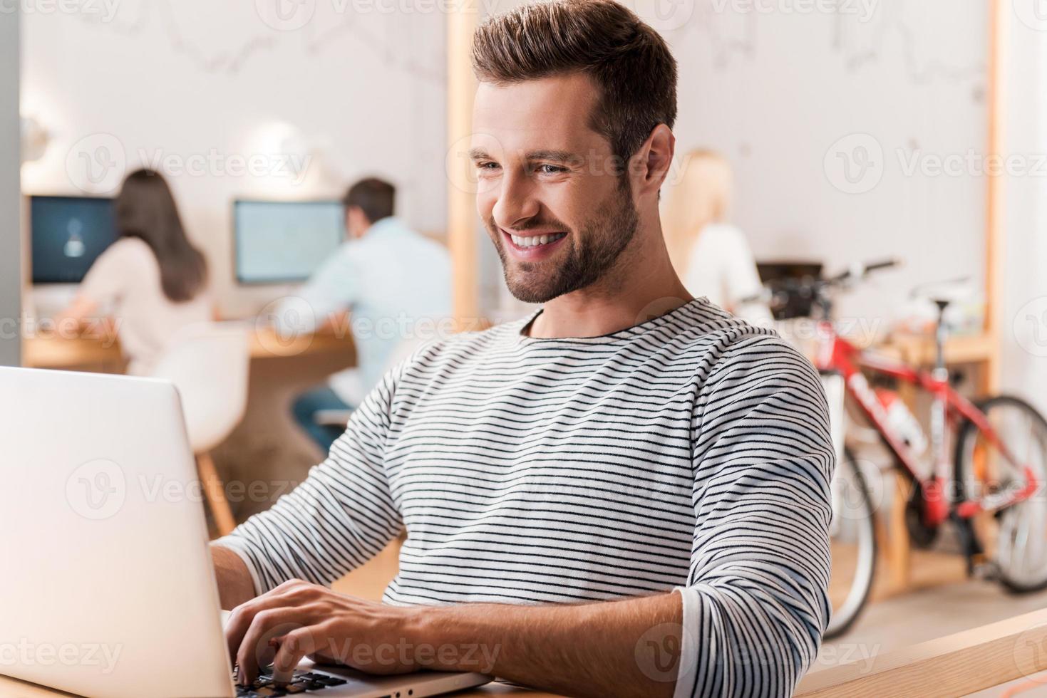trabajando con placer. un joven apuesto que trabaja en una laptop y sonríe mientras sus colegas trabajan en segundo plano foto