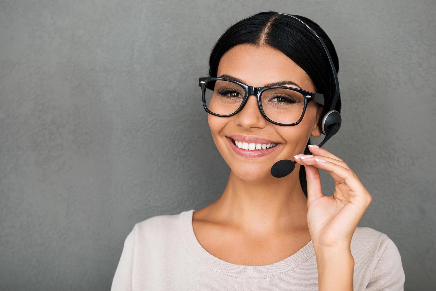 Service with happy smile. Cheerful young female customer service looking at camera and smiling while standing against grey background photo