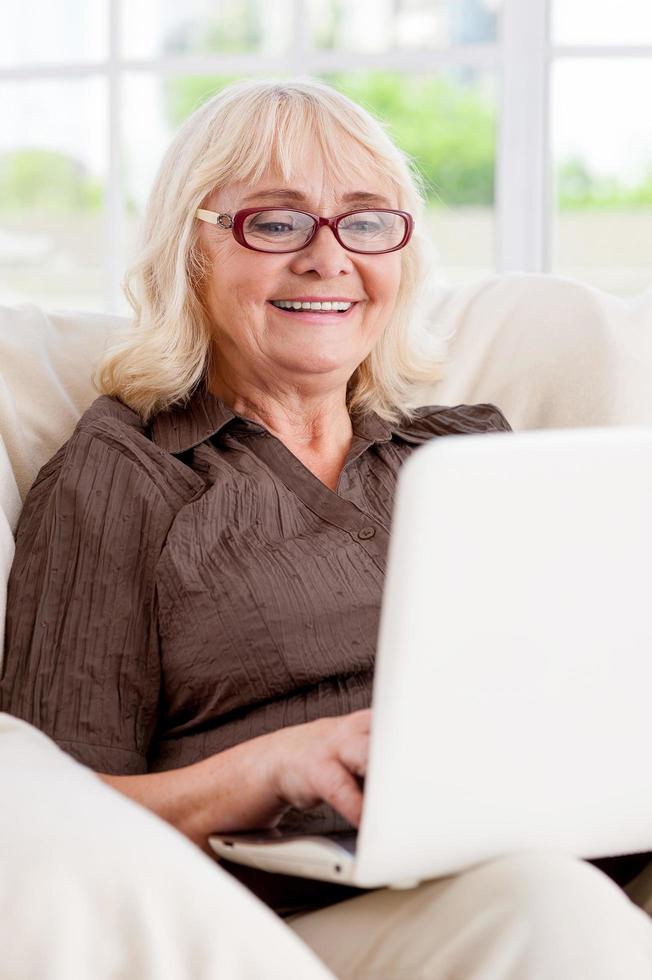 navegar por la red es divertido. anciana trabajando en una laptop y sonriendo mientras se sienta en la silla foto