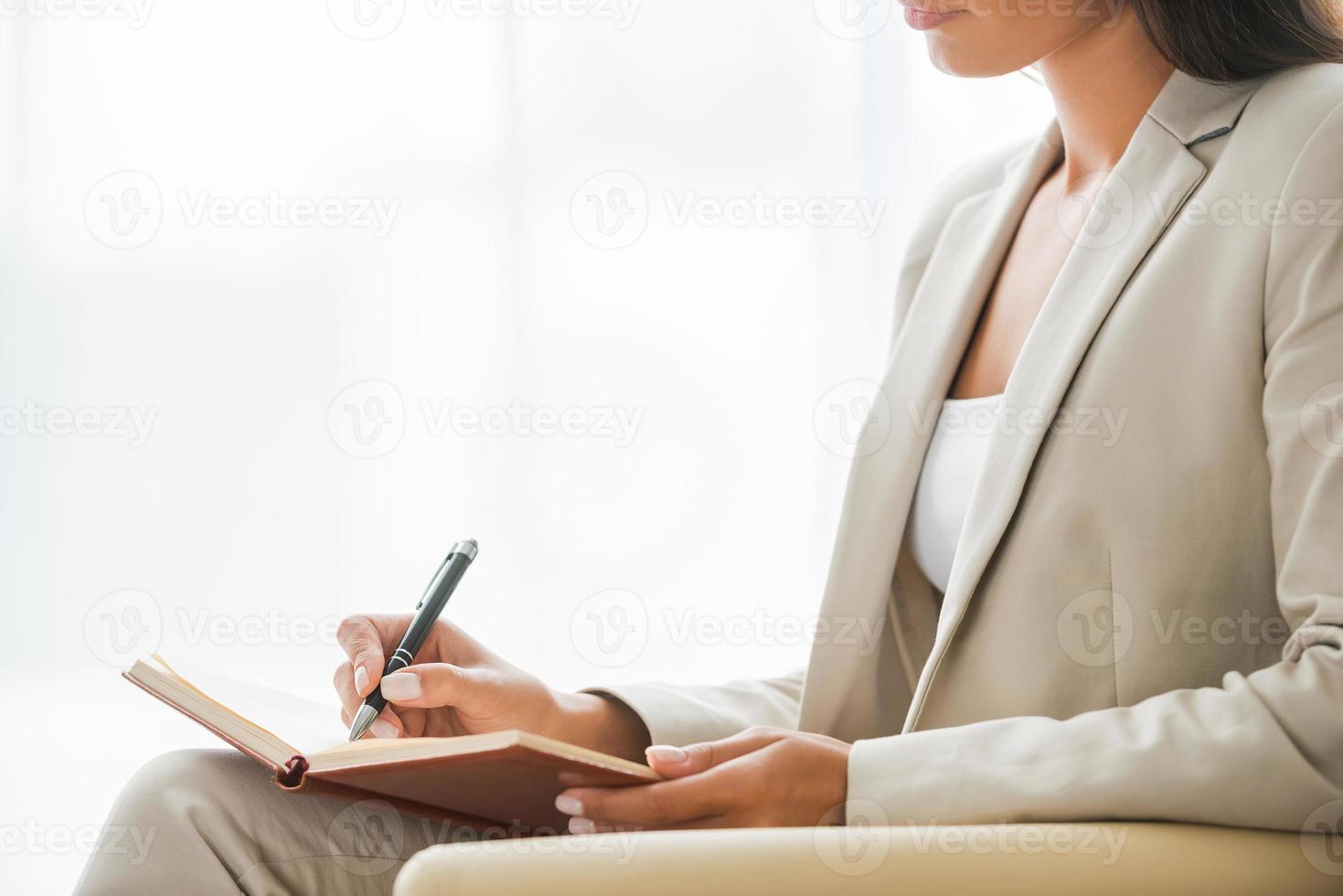 Making business notes. Side view of young businesswoman in suit writing something in note pad while sitting at the chair photo