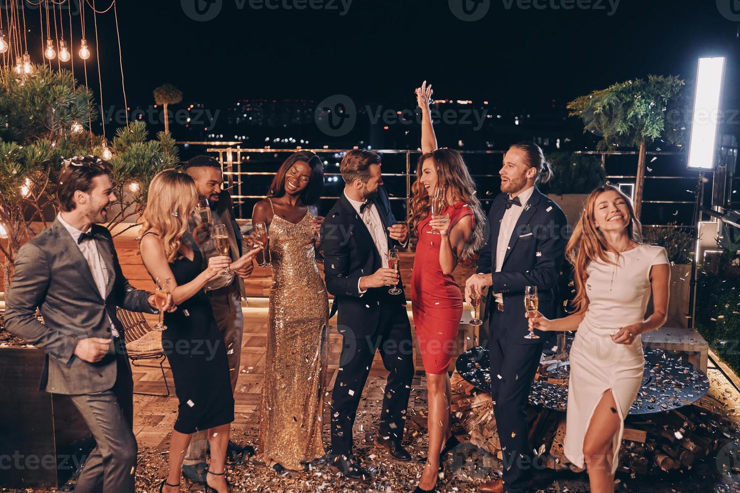 Group of happy people in formalwear dancing and having fun together with confetti flying all around photo