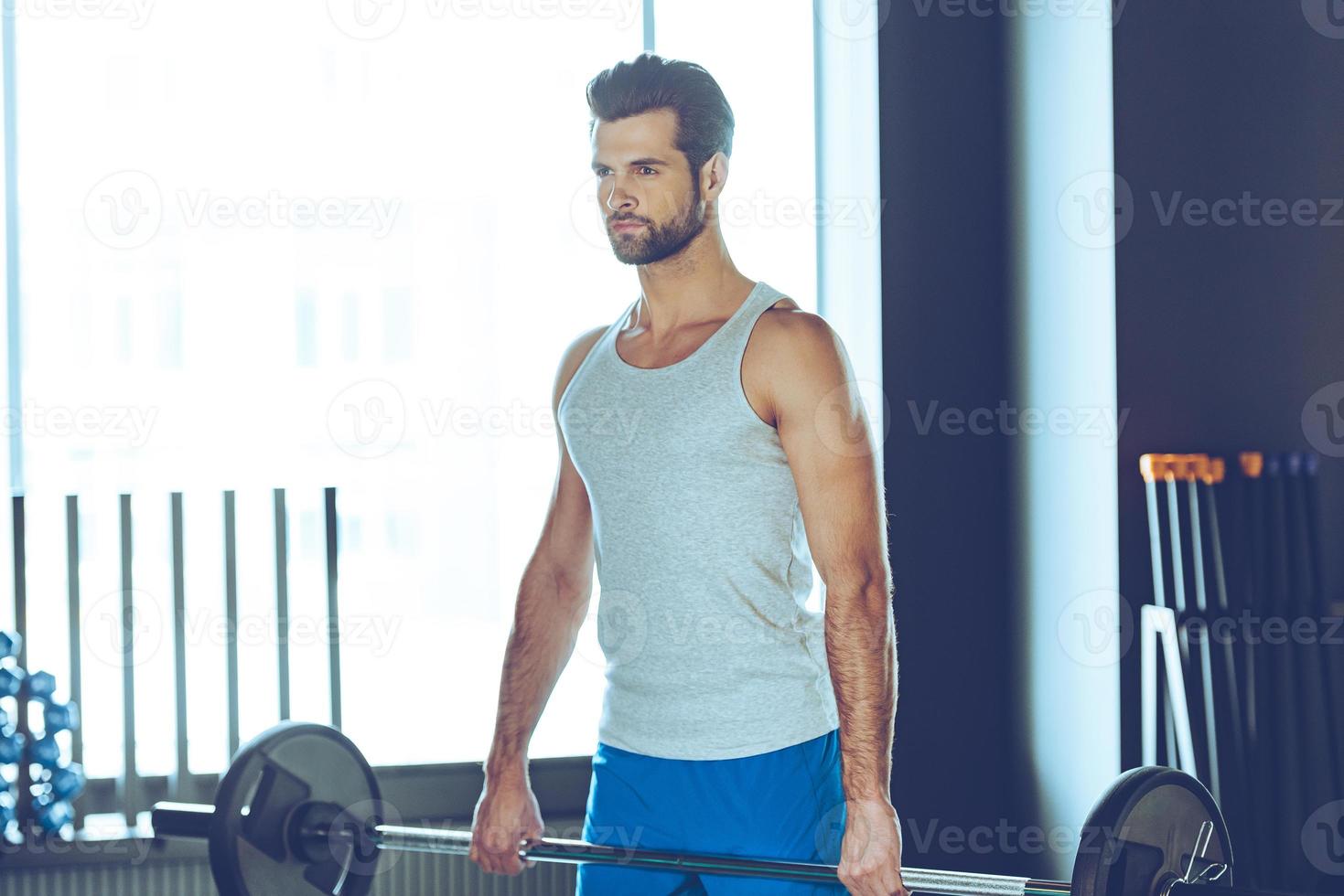 Concentrated athlete. Young handsome man in sportswear lifting barbell at gym photo