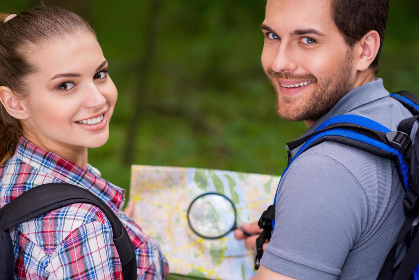 Happy tourist couple. Beautiful young loving couple carrying backpacks and looking over shoulder with smile while walking along the forest pathway photo