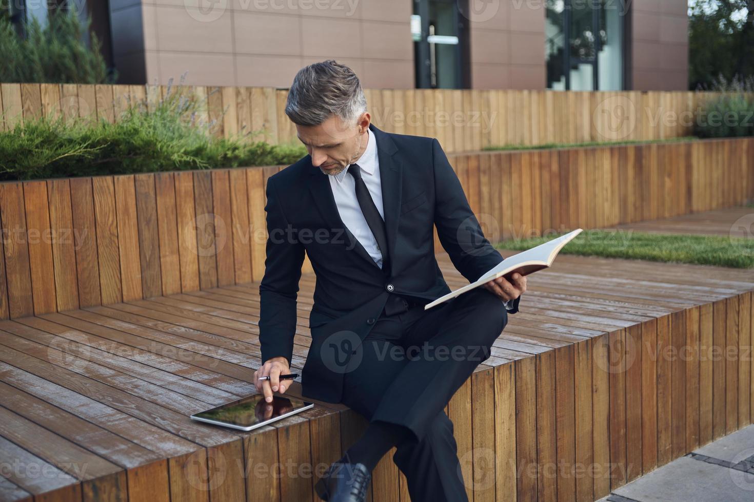 Confident mature businessman using digital tablet and making notes while sitting outdoors near office building photo