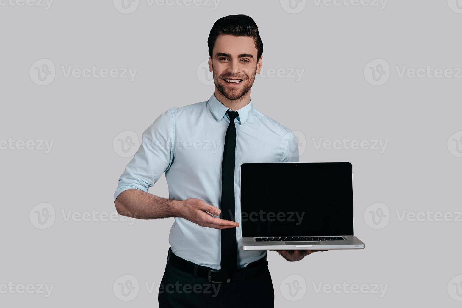 Copy space on his laptop.  Good looking young man in white shirt and tie pointing copy space on his laptop and smiling while standing against grey background photo