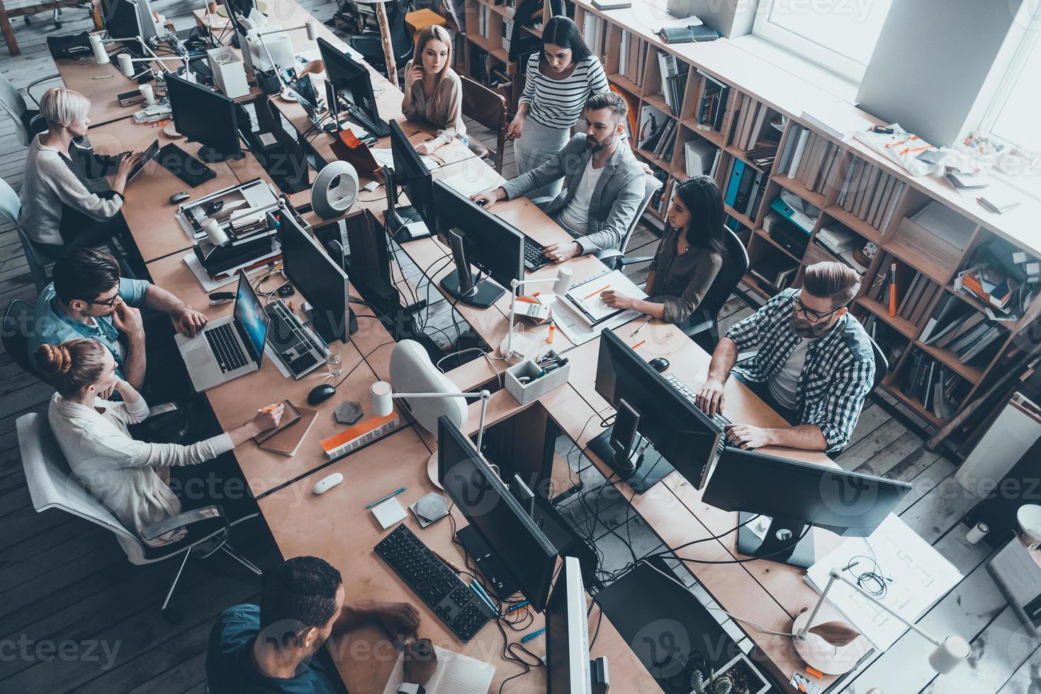 Confident business experts a work. Top view of group of young business people in smart casual wear working together while sitting at the large office desk photo