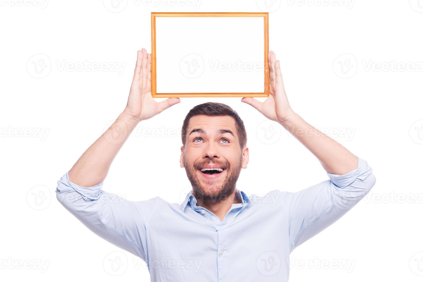 Happy about that... Handsome young man in shirt holding a picture frame upon his head and looking up on it while standing against white background photo