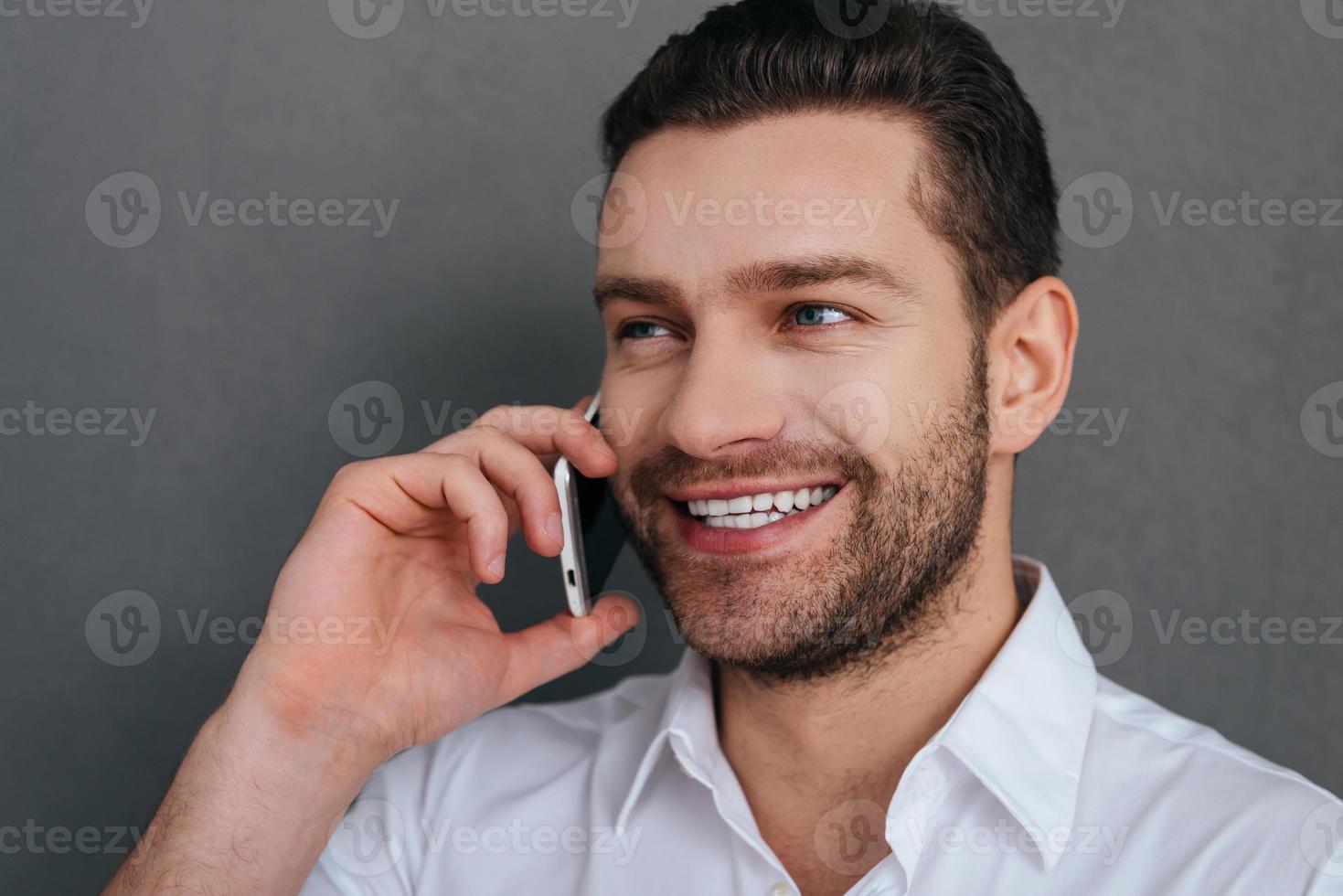 Good talk. Handsome young man talking on the mobile phone and smiling while standing against grey background photo