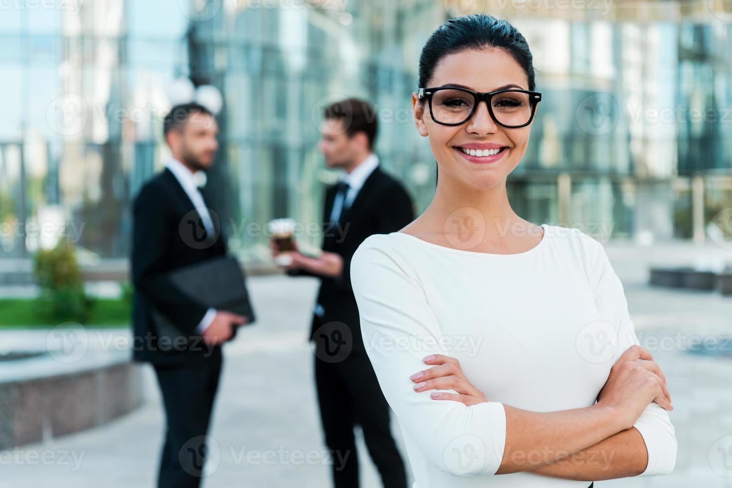 Confident businesswoman. Smiling young businesswoman keeping arms crossed and looking at camera while two her male colleagues talking to each other in the background photo
