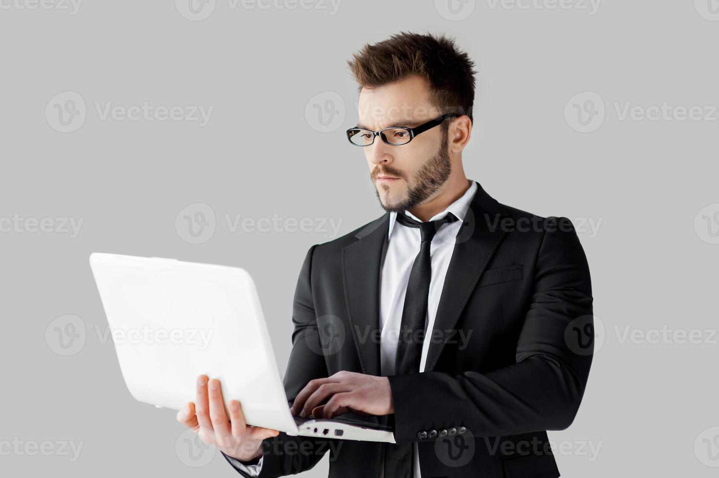 Businessman surfing the net. Cheerful young man in formalwear and glasses working on laptop while standing against grey background photo