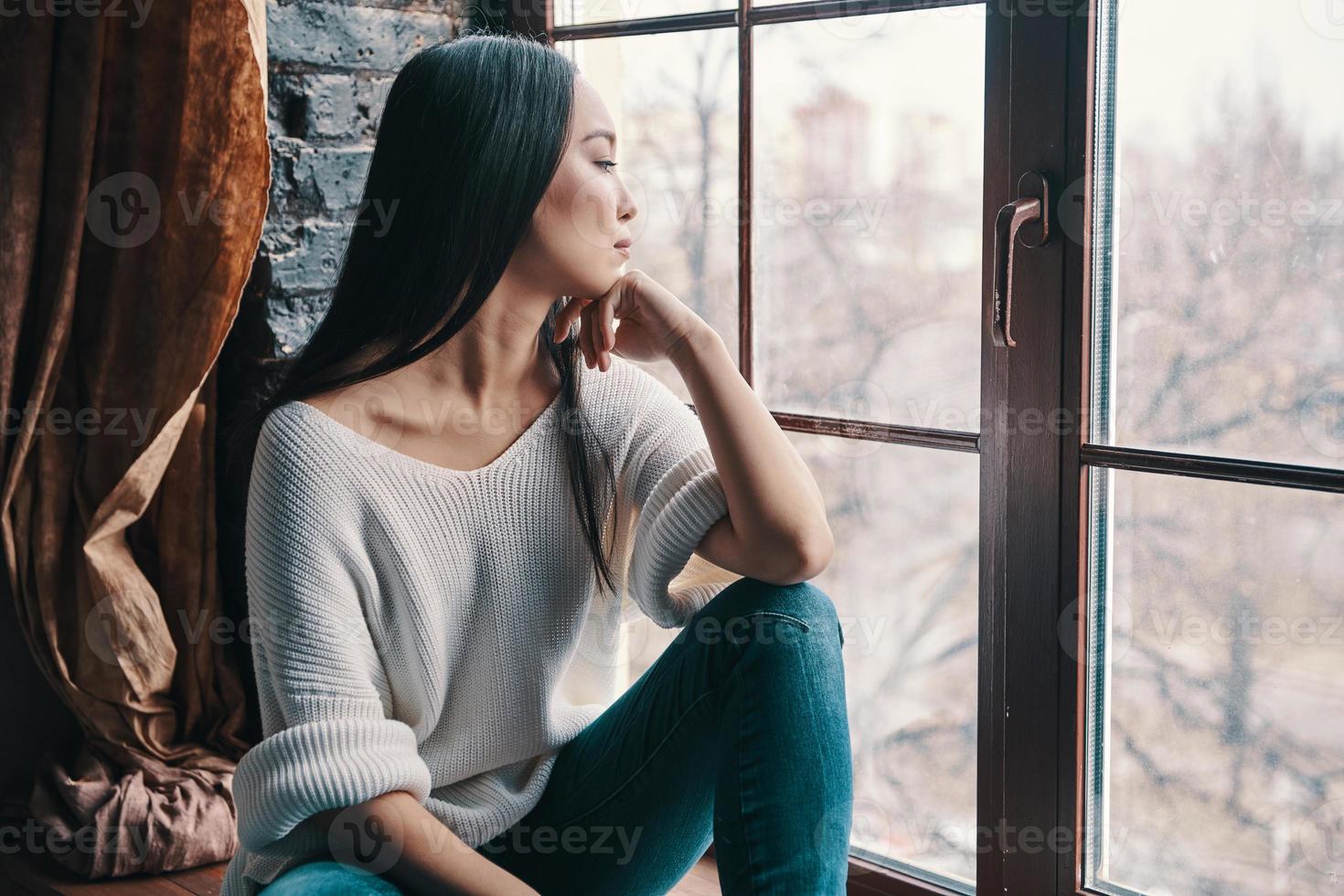 Thinking about.... Attractive young woman keeping hand on chin and looking away while sitting on the window sill at home photo