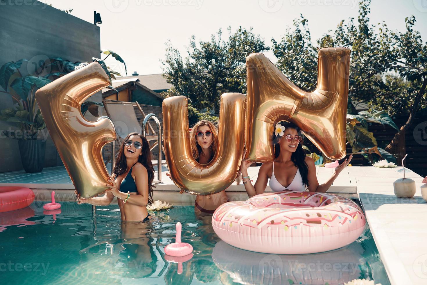 Attractive young women in swimwear smiling and lifting up balloons while standing in the pool outdoors photo