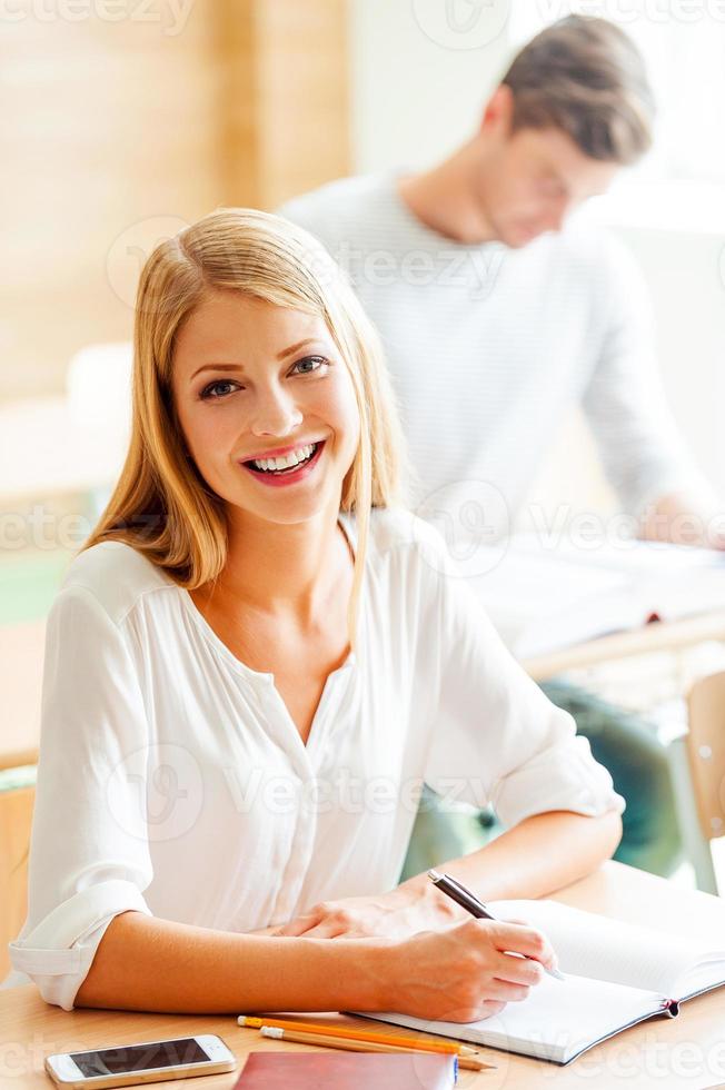 Developing good study habits. Joyful young woman writing in note pad and smiling while sitting at her desk in the classroom photo