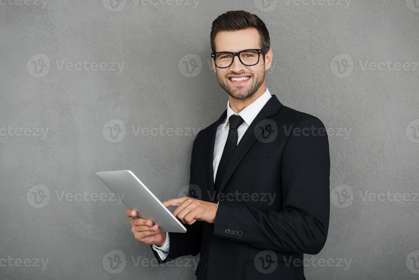 Doing his business in easy way. Happy young businessman holding digital tablet and looking at camera while standing against grey background photo