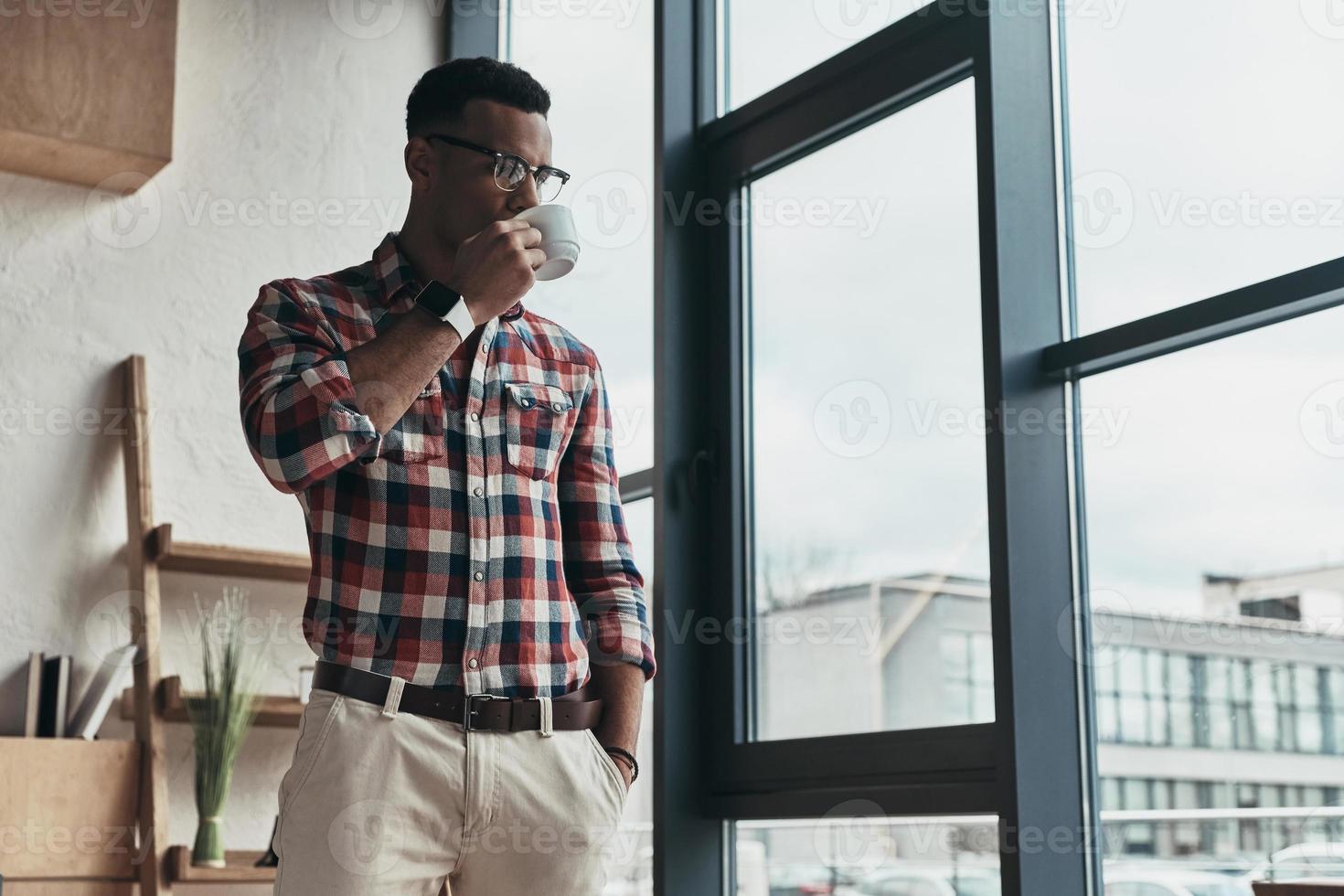 On a little break. Handsome young man drinking coffee and keeping hand in pocket while standing near the window photo