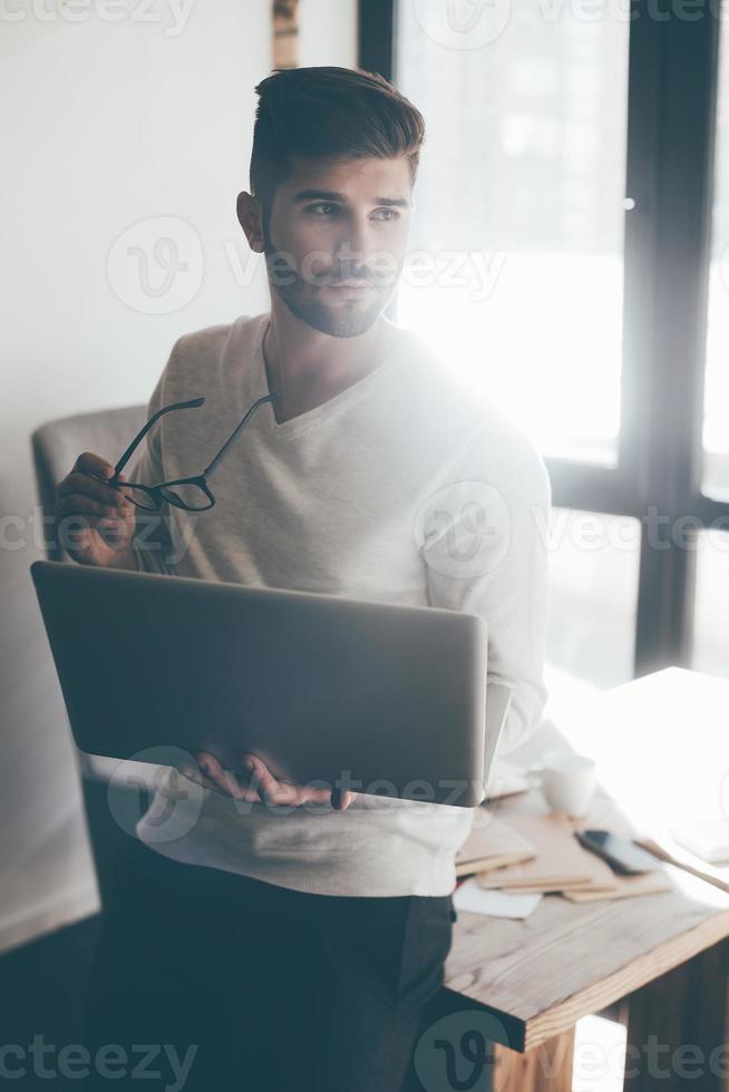 Thinking about solution. Thoughtful young man carrying laptop and eyeglasses while leaning at the desk in office and looking away photo