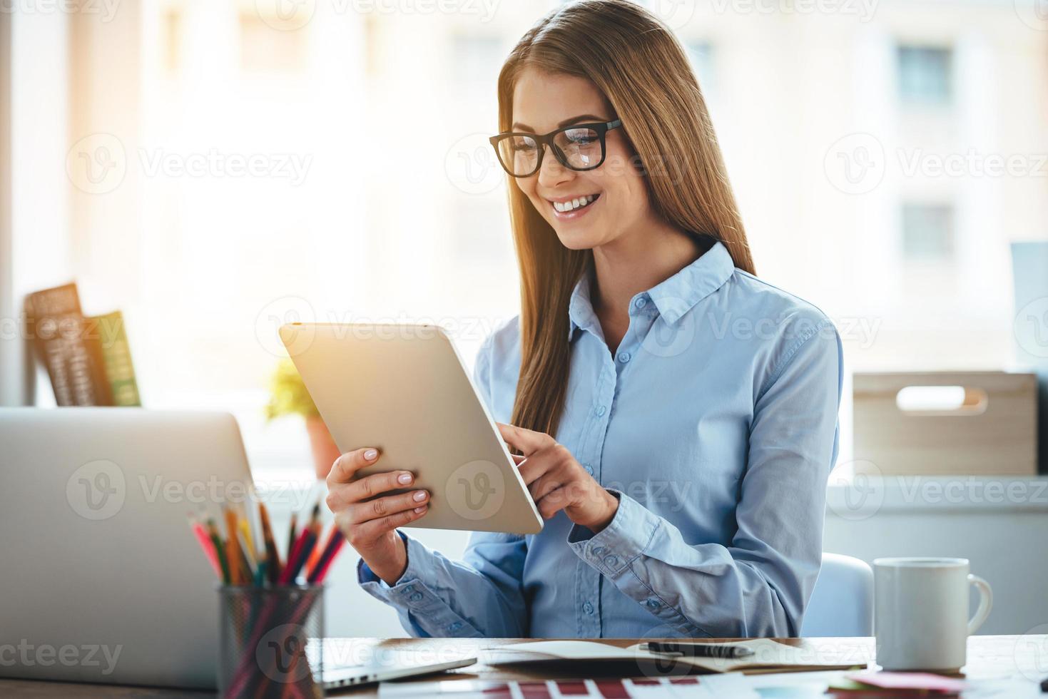 Lets check my timetable Cheerful young beautiful woman in glasses using her touchpad with smile while sitting at her working place photo