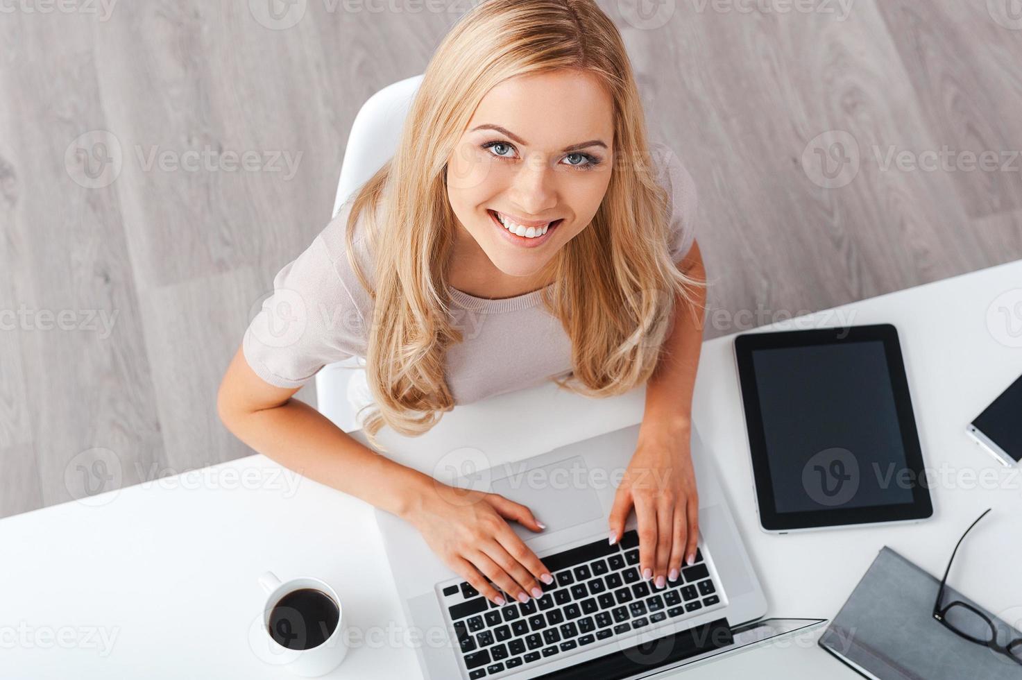 Enjoying productive day. Top view of cheerful young businesswoman working on laptop and looking at camera while sitting at her working place photo