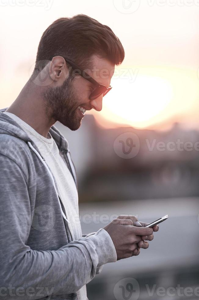 Texting to friend. Side view of handsome young man in sunglasses holding mobile phone and smiling while standing outdoors photo