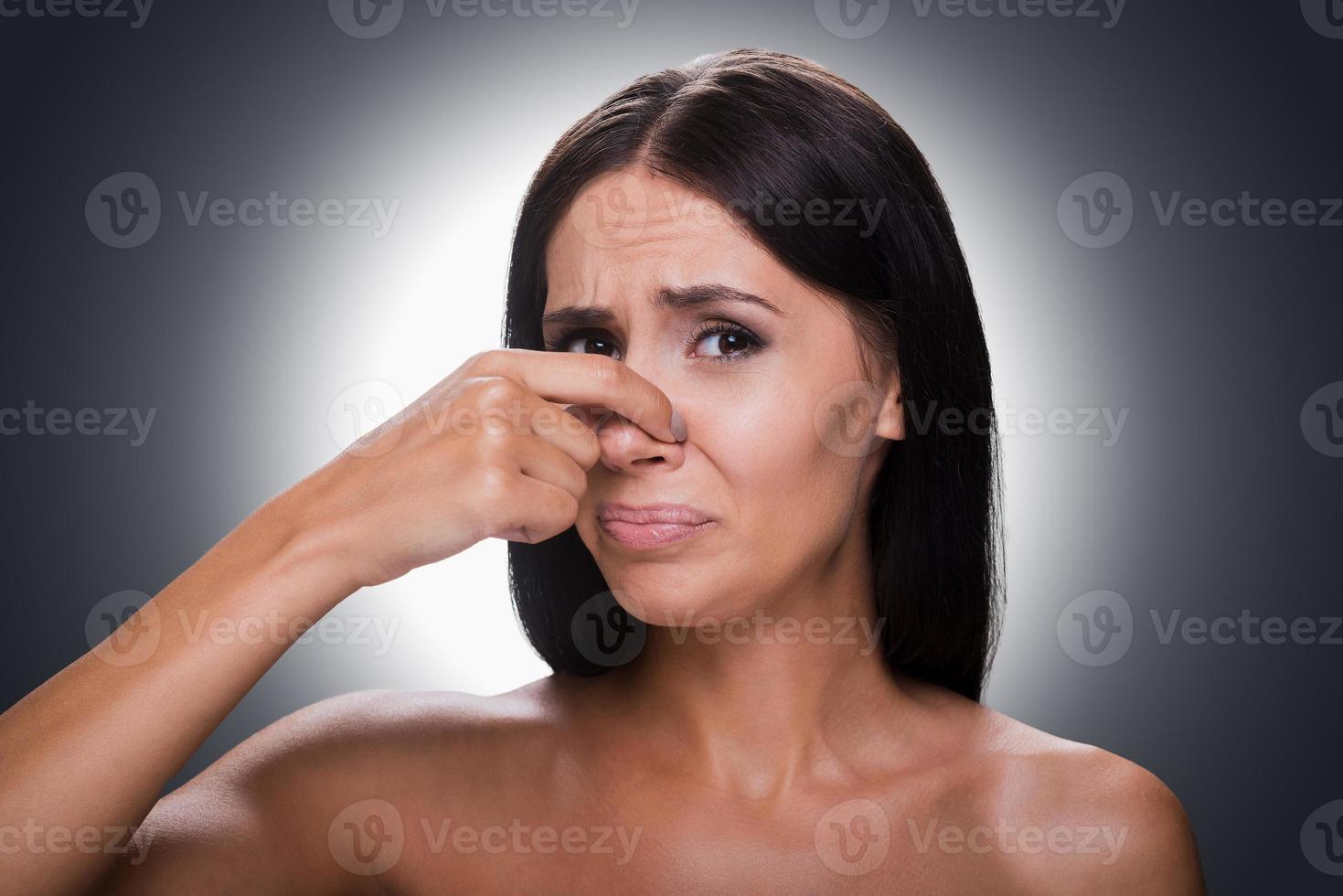 Unpleasant smell. Portrait of frustrated young shirtless woman holding fingers on nose and making a face while standing against grey background photo