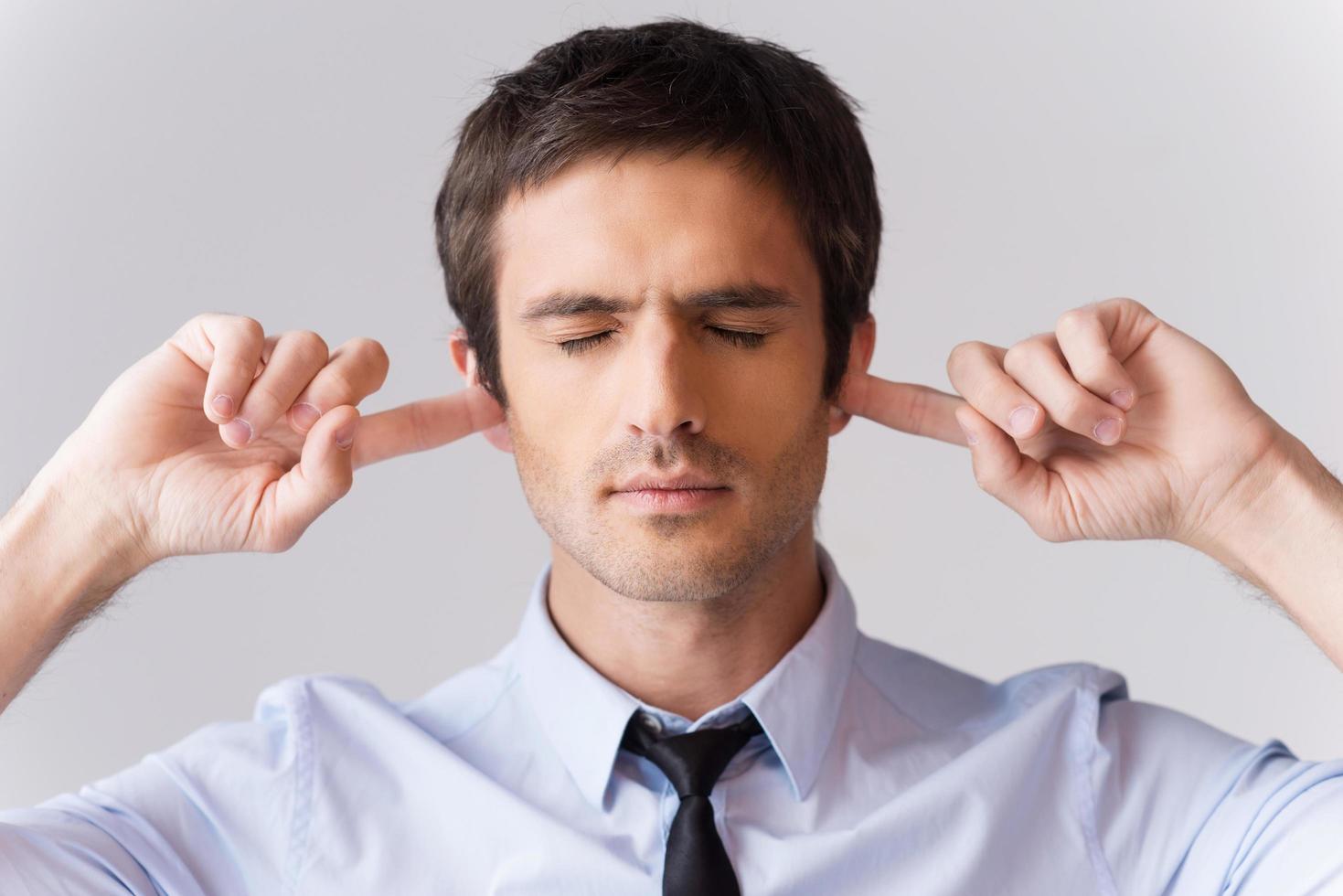 Too loud for me. Handsome young man in shirt and tie holding fingers in ears and keeping eyes closed while standing against grey background photo