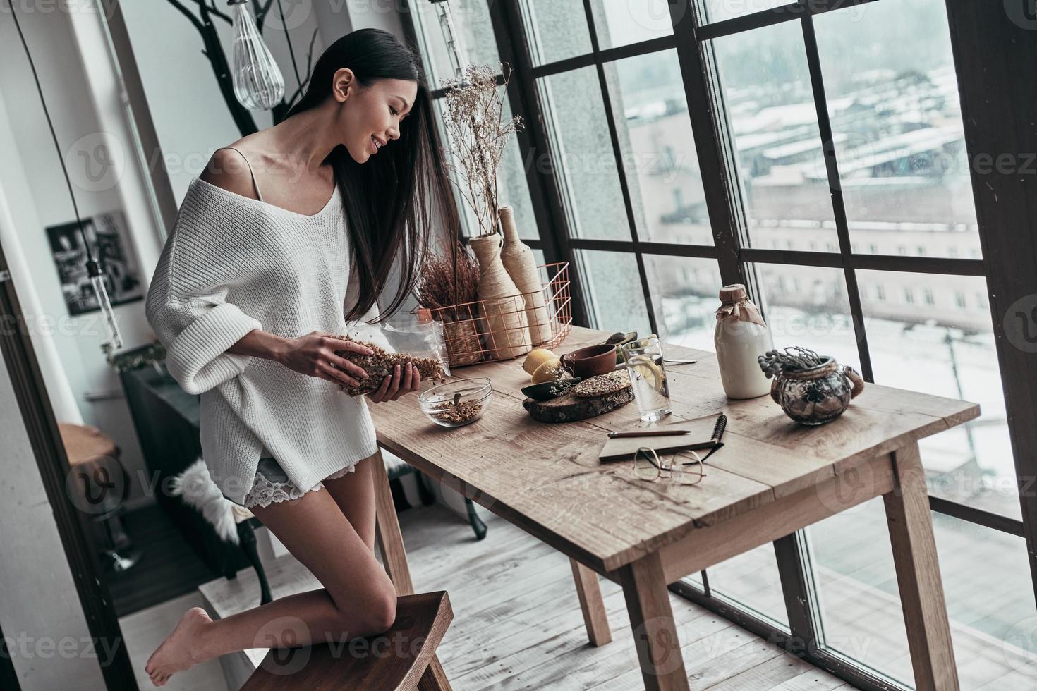Delicious breakfast. Top view of attractive young woman pouring cereal and smiling while standing near the window at home photo
