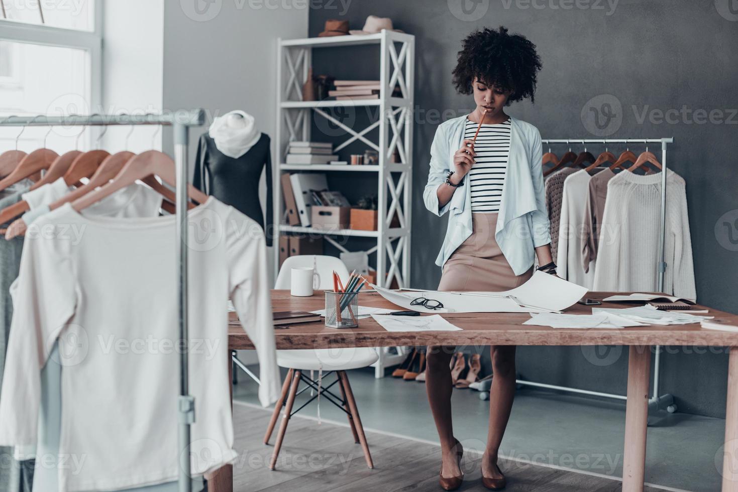 Generating fresh ideas. Full length of young African woman touching lips with pencil and looking at paper while standing in her workshop near the clothes hanging on the racks photo