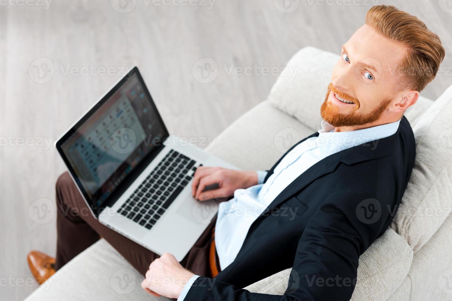 Working with pleasure. Top view of happy bearded businessman working on laptop and smiling at camera while sitting on sofa photo