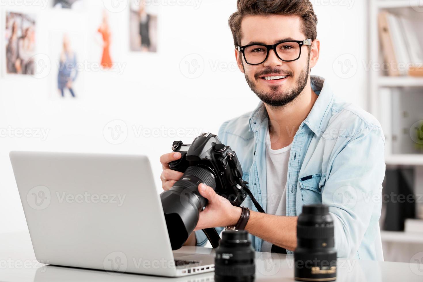 amo mi trabajo joven feliz sosteniendo la cámara y sonriendo mientras está sentado en su lugar de trabajo foto