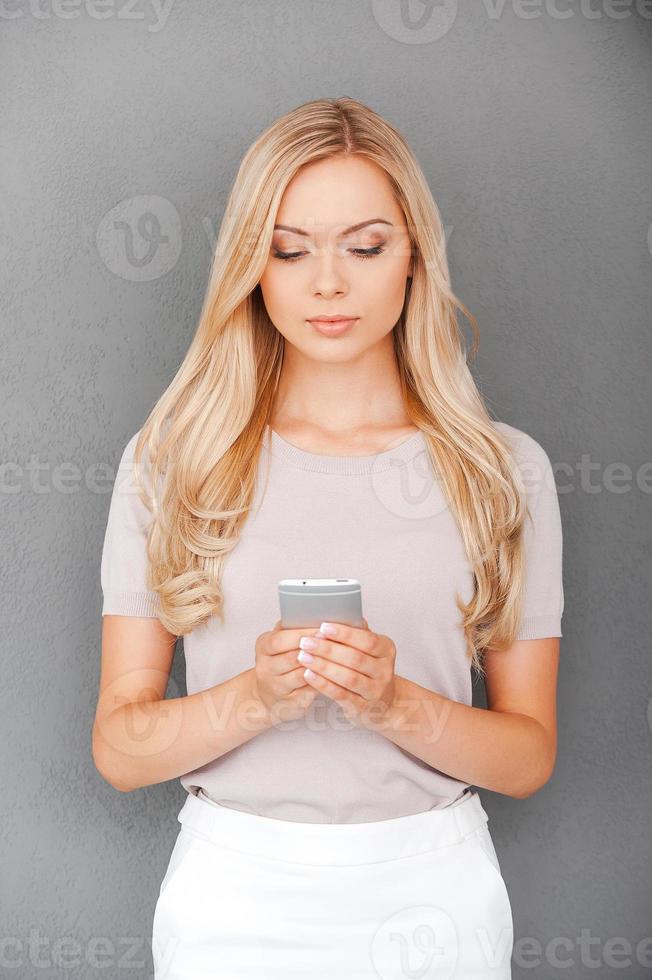 Checking her day schedule. Confident young blond hair woman holding mobile phone while standing against grey background photo