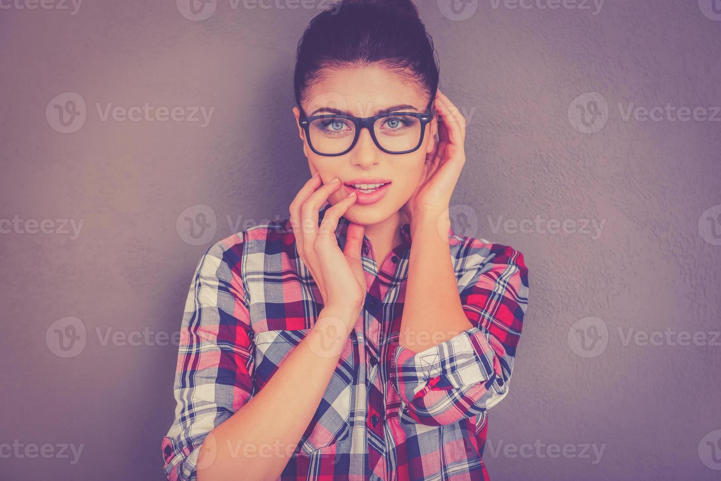 Feeling flirty. Beautiful young woman in eyeglasses holding finger in mouth and looking at camera while standing against grey background photo