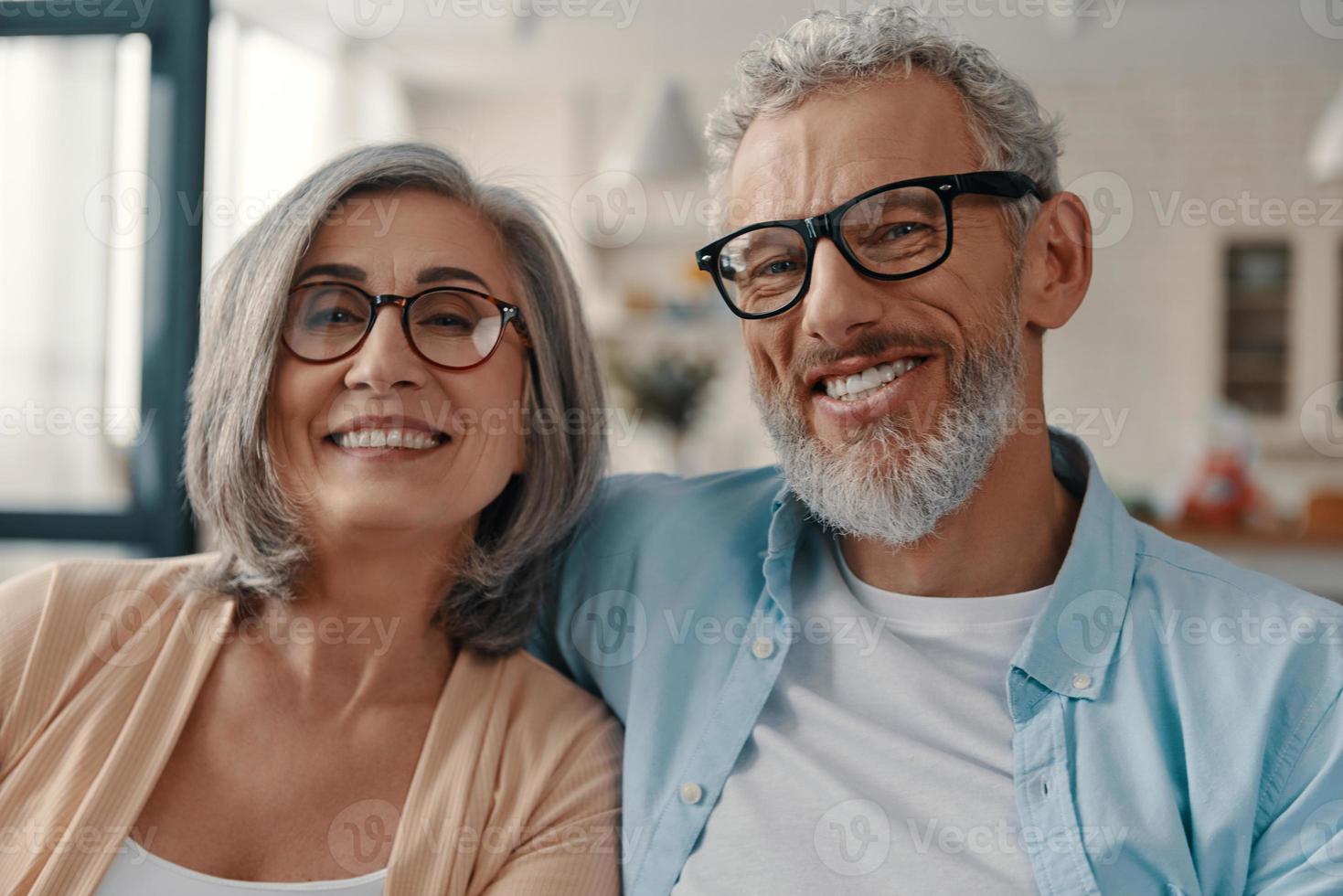 Beautiful senior couple in casual clothing smiling and looking at camera while spending time at home photo