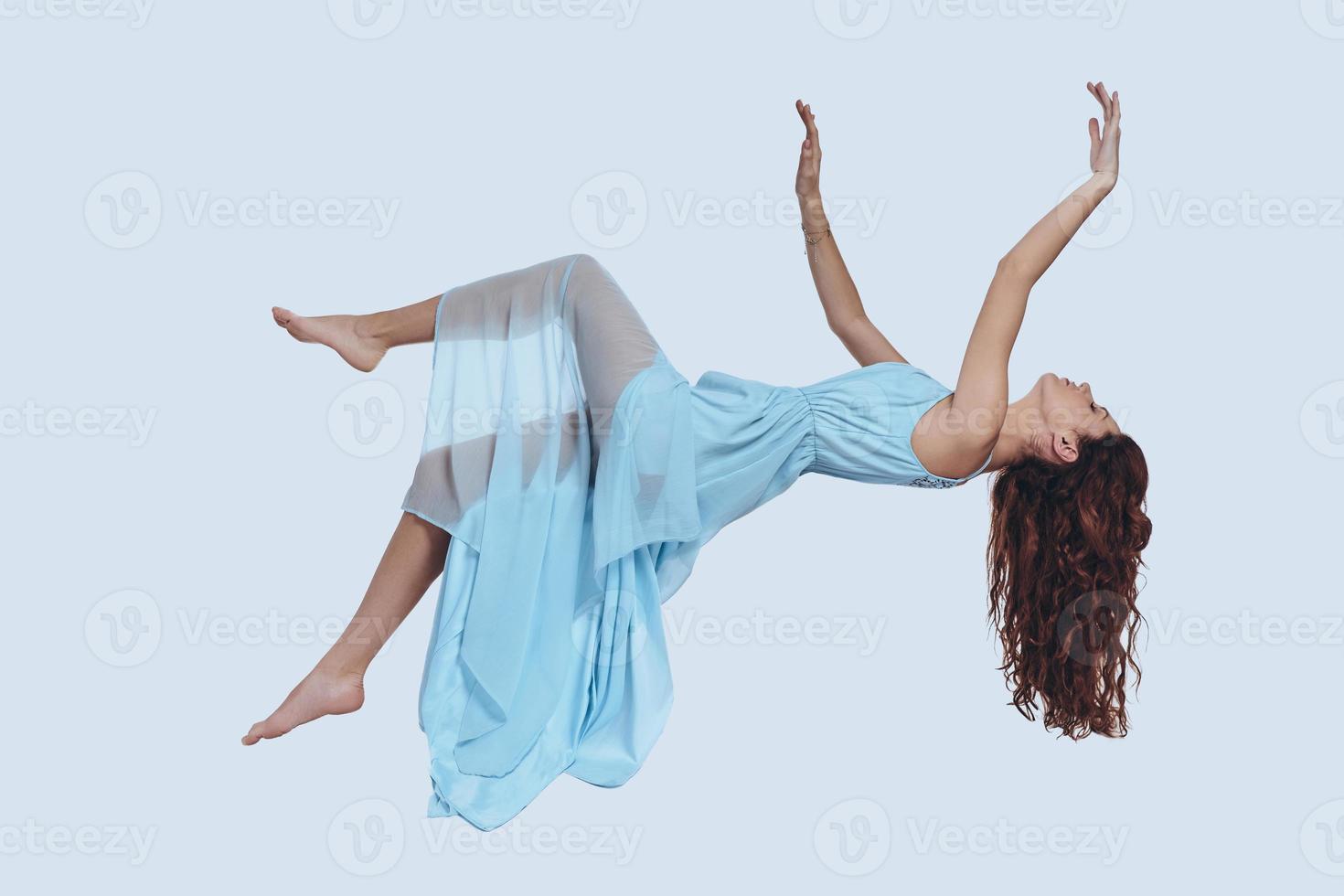 Flying so high. Studio shot of attractive young woman in elegant dress keeping arms outstretched while hovering against grey background photo